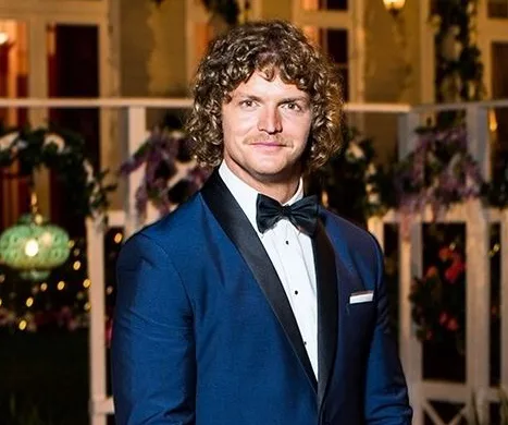 Man in a blue tuxedo and bow tie standing outdoors at night with floral decorations in the background.