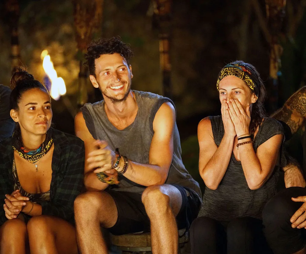 Three "Survivor" contestants sitting, one smiling, two appearing anxious, at tribal council with torches in the background.