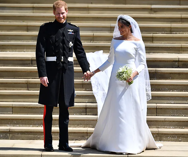 Prince in military uniform and bride in white dress stand hand in hand on steps, smiling.