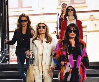 Four women in stylish outfits and sunglasses walk down stairs confidently in an urban setting.
