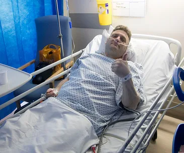 Man in hospital bed giving a thumbs-up, wearing a nasal cannula and hospital gown.