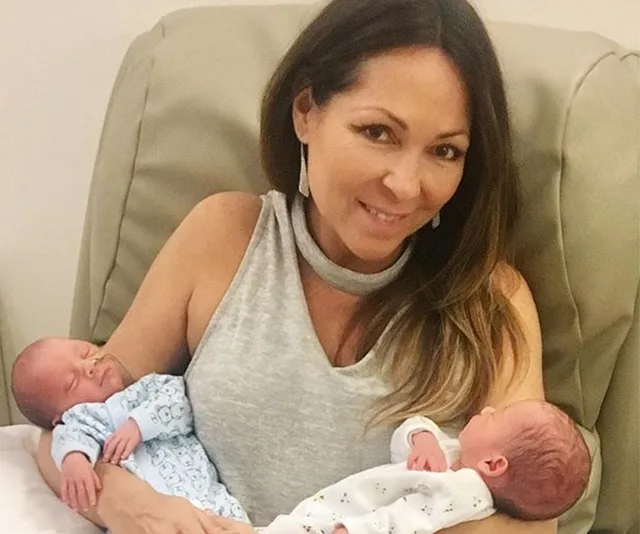 Woman smiling while holding two newborn twins in her arms, sitting on a beige chair.