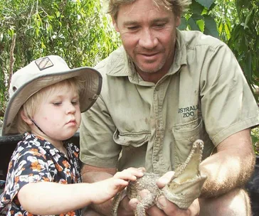 Father and child gently holding a baby crocodile at a zoo, surrounded by lush greenery.