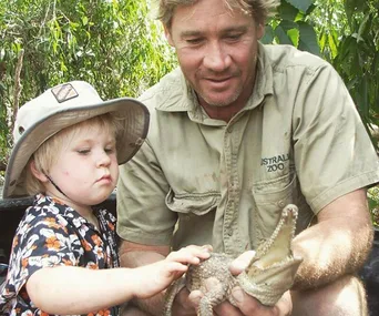 Father and child gently holding a baby crocodile at a zoo, surrounded by lush greenery.