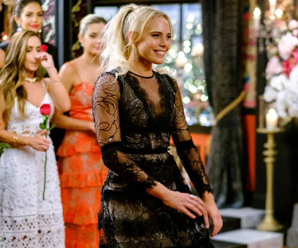 A contestant on "The Bachelor Australia" smiles, walking in a room with others in elegant dresses holding roses in the background.
