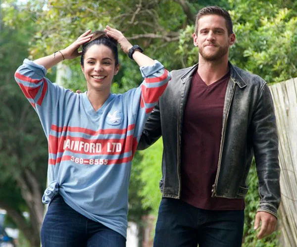 A woman in a sports jersey and a man in a leather jacket walk outdoors, both smiling, with trees in the background.