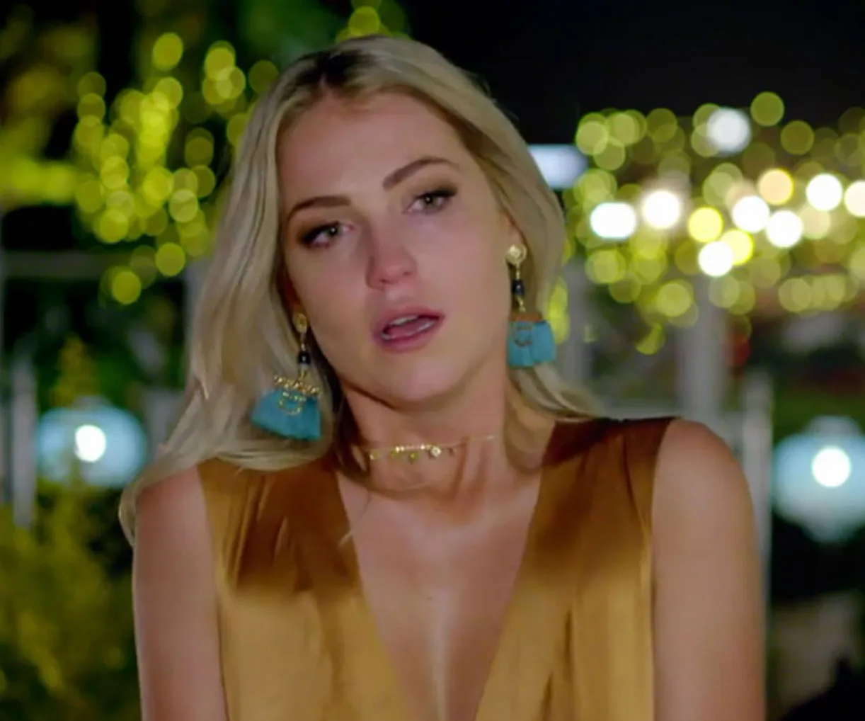 A woman with blonde hair looks emotional, wearing statement earrings and a gold dress, with blurred lights in the background.