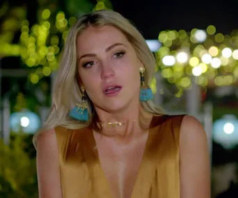 A woman with blonde hair looks emotional, wearing statement earrings and a gold dress, with blurred lights in the background.