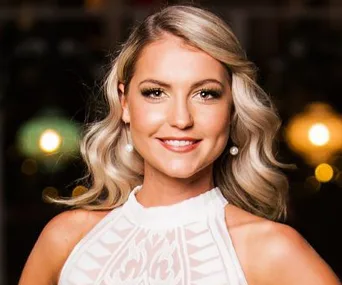 A woman with wavy blonde hair in a white dress smiles against a blurred background with warm lights.