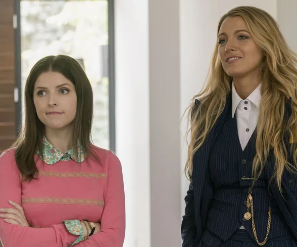 Two women, likely characters from "A Simple Favor," stand indoors; one in a pink sweater, the other in a pinstripe suit.