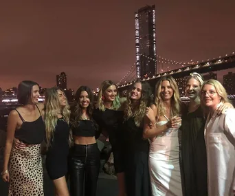 Group of women posing at night by a lit-up bridge, with city skyline in the background.