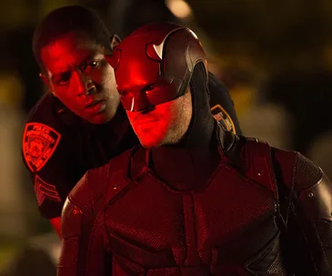 Daredevil in red lighting, wearing his suit and mask, stands with a concerned police officer behind him.