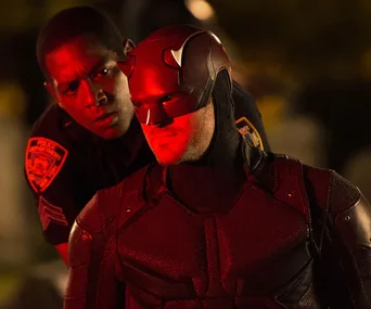 Daredevil in red lighting, wearing his suit and mask, stands with a concerned police officer behind him.