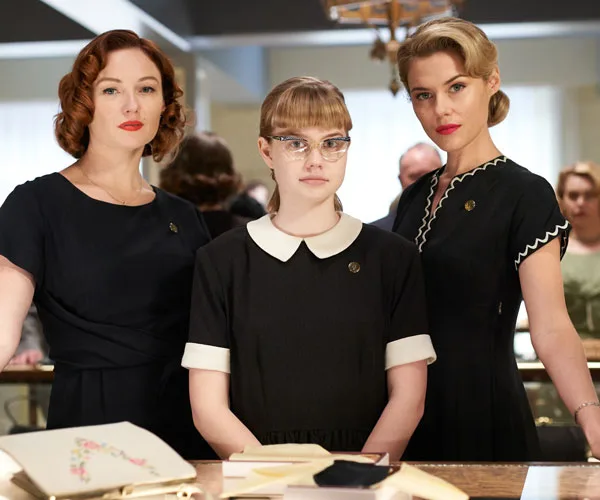 Three women in vintage 1950s black dresses stand together, the middle one wearing glasses. Scene from "Ladies in Black."