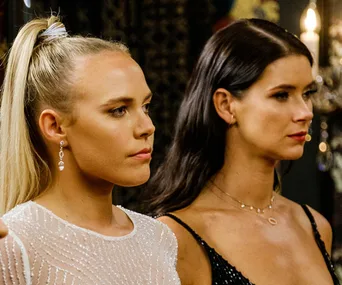 Two women stand side by side in evening dresses, focused intently on something off-camera.