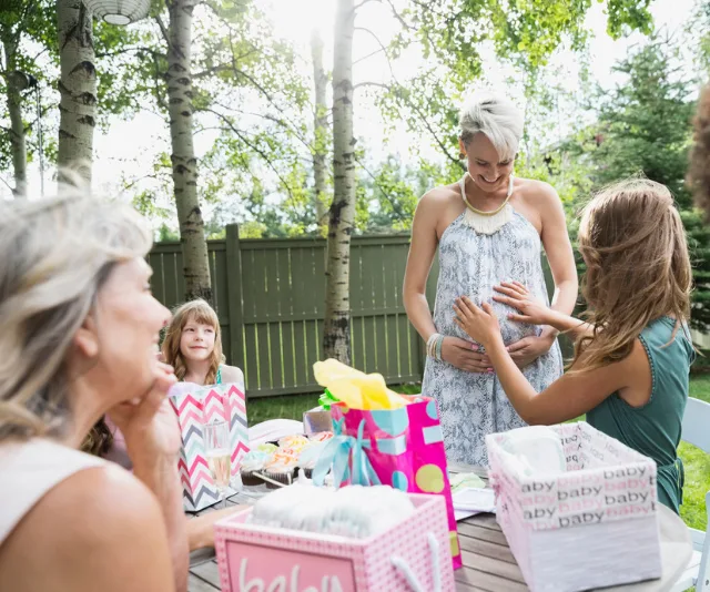 Pregnant woman at outdoor baby shower, surrounded by gifts and guests touching her belly.