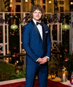 A man in a blue tuxedo stands outside a romantically lit setting with candles and flowers.