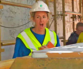 Person in a construction setting, wearing a hard hat and hi-vis vest, holding a notebook, appears to be speaking energetically.
