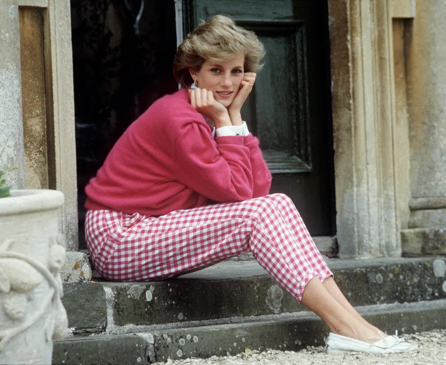 A woman sitting on steps, wearing a pink sweater and checkered pants, with her hands under her chin, smiling gently.
