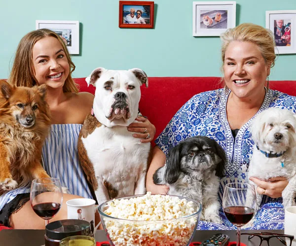 Two women sit on a sofa with four dogs, popcorn, and wine, smiling for the camera in a cozy living room setting.
