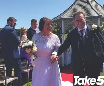An older couple in wedding attire joyfully walking down the aisle, surrounded by guests and a gazebo in the background.