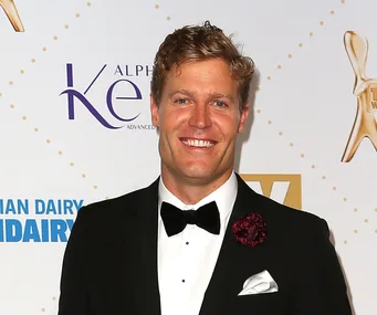 Man in a black tuxedo with a bow tie smiles in front of a backdrop with logos at an event.