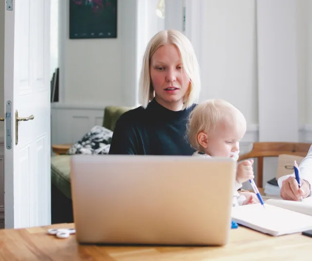 A woman working on a laptop at a table with a child sitting beside her, holding a pen over a notebook.