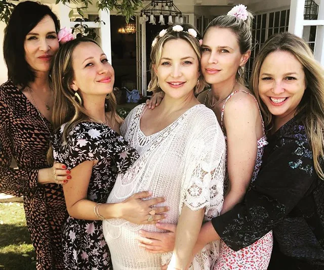 Five women, one pregnant, smiling and embracing at a baby shower, wearing floral outfits and flower crowns in a garden.