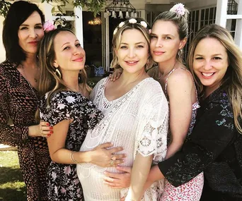 Five women, one pregnant, smiling and embracing at a baby shower, wearing floral outfits and flower crowns in a garden.