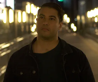 A person stands on a dimly lit city street at night, with blurred lights in the background.