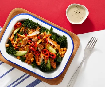 A colorful salad with shredded chicken, chickpeas, kale, cucumber, and chili peppers, served with a creamy dressing.