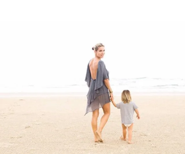 Mother and child walk hand in hand on a sandy beach, with the ocean in the background.