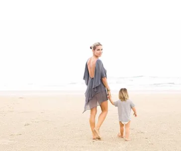 Mother and child walk hand in hand on a sandy beach, with the ocean in the background.