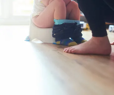 Toddler sitting on a potty chair with their pants down, while an adult stands nearby on a wooden floor.