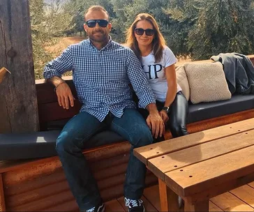 Couple sitting outdoors on a bench, both wearing sunglasses, surrounded by cushions with a wooden table in front.