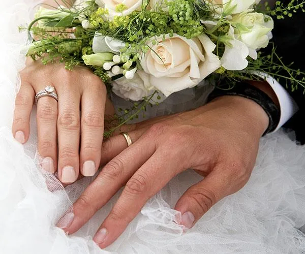 Close-up of newlywed couple's hands with wedding rings, holding a bouquet of white roses and greenery.