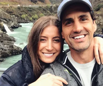 Couple smiling by a scenic river backdrop, wearing casual outdoor attire.