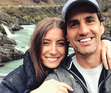 Couple smiling by a scenic river backdrop, wearing casual outdoor attire.