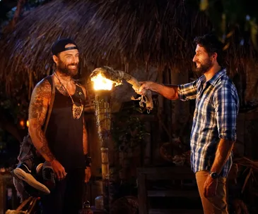 Two men standing near a burning torch in a tribal setting, one passing a torch to the other with a smile.