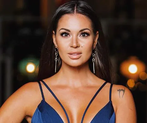 Dasha from "The Bachelor" poses in a blue dress with earrings, against a blurred background with lights.