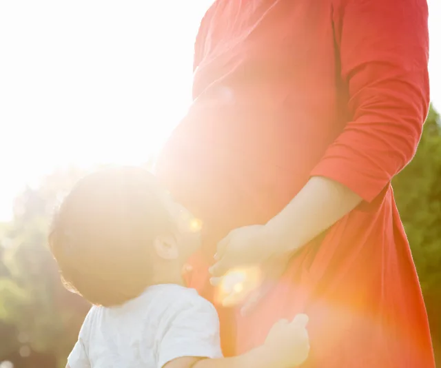 Pregnant woman in red dress with child outdoors, sunlight in background.