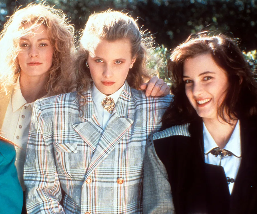 Three young women pose together, dressed in 1980s fashion, featuring blazers and bold hairstyles, outdoors.