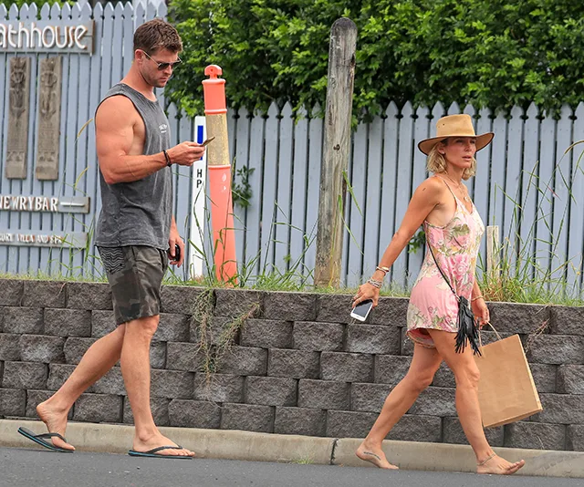 Man and woman walking barefoot on a street, both holding phones; man in tank top, woman in a dress and hat.