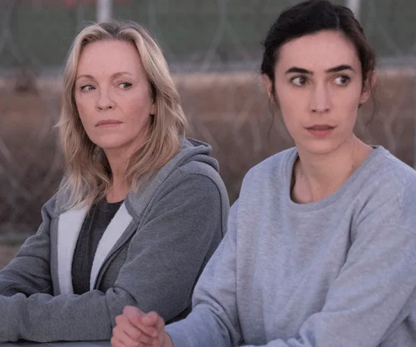 Two women sit outdoors in casual clothing, looking in different directions with serious expressions.