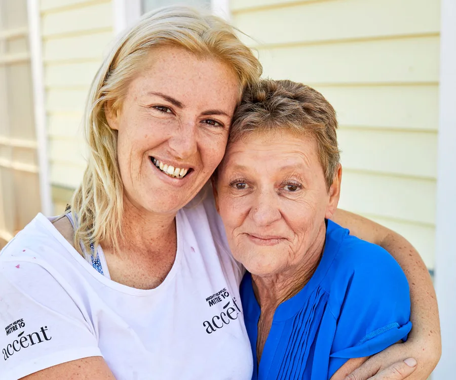 Two women embracing and smiling outside a yellow house. One is wearing a white "accent" shirt, the other a blue top.