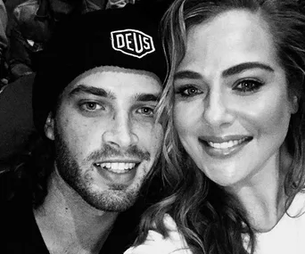 Two people taking a smiling selfie, one wearing a black beanie with "DEUS" written on it. Black and white photo.