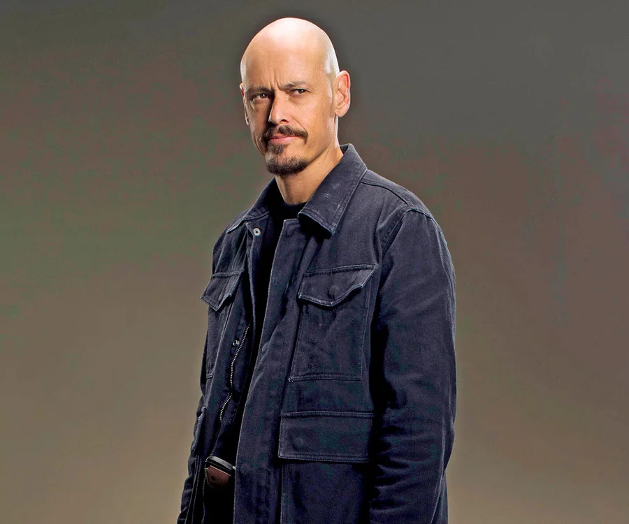 Man in a dark jacket, posing with a determined expression against a neutral background.