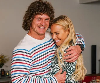 A man in a striped shirt and a woman in a patterned dress embrace, smiling warmly indoors.