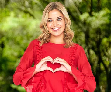 Woman smiling in a red dress, making a heart shape with her hands in front of a leafy background.