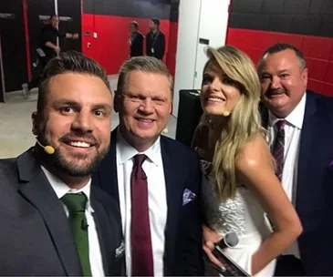 Four presenters with microphones pose for a selfie backstage, smiling at the camera in formal attire.